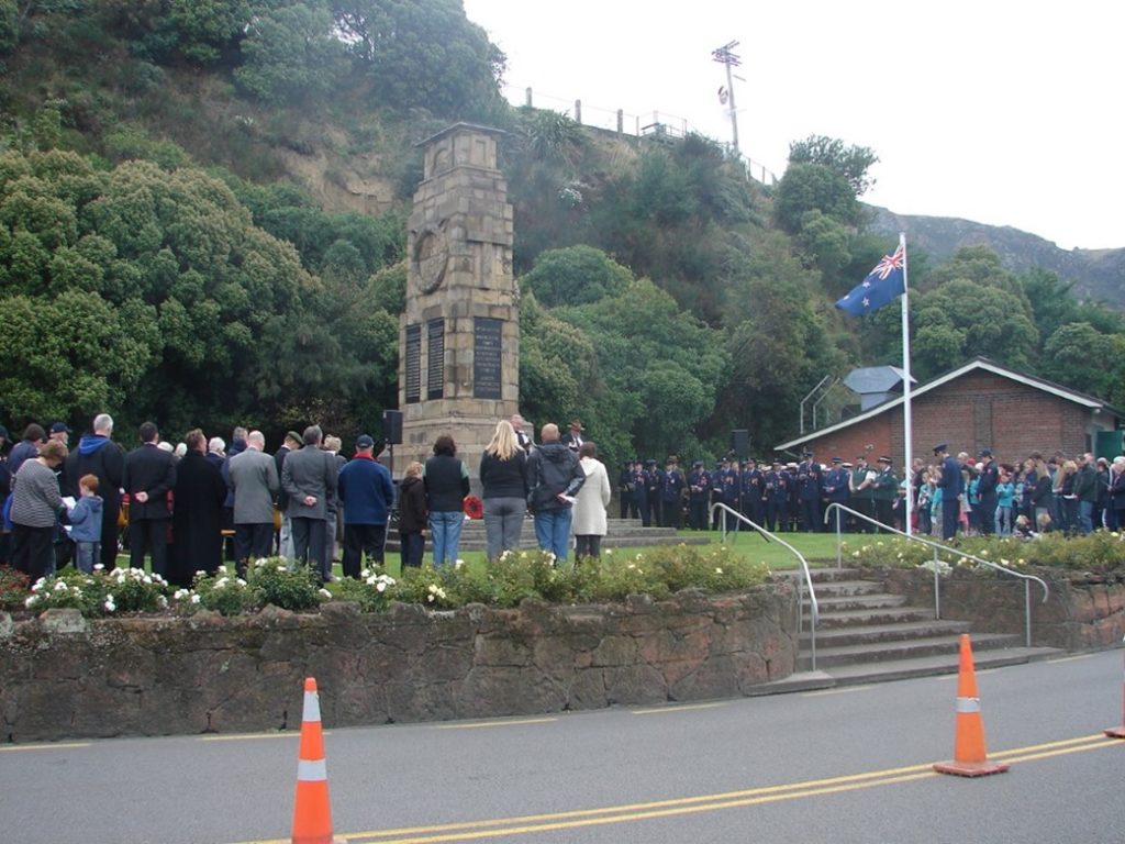 Photo taken across the road at the Lyttelton War Memorial showing people partaking in the service with the NZ ensign flying